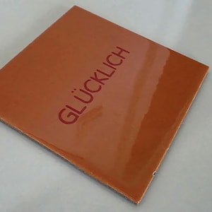 Könnte beinhalten: Eine quadratische, glänzende, orange-braune Keramikfliese mit dem Wort GLÜCKLICH, das in einer dunkelroten, serifenlosen Schrift aufgedruckt ist. Die Fliese hat eine glatte, reflektierende Oberfläche und scharfe, saubere Kanten.