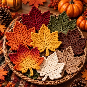 May include: A collection of crocheted maple leaf coasters in various autumn colors including deep red, forest green, golden yellow, burnt orange, dark brown, cream, and tan, all arranged together inside a round woven wicker basket.