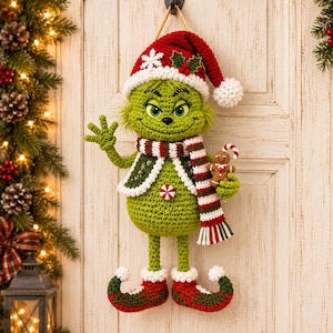 Pode incluir: Um enfeite de parede de crochê em forma de criatura verde usando um gorro de Papai Noel vermelho e branco com um floco de neve, um cachecol listrado e um colete verde. Segura um pequeno biscoito de gengibre, um doce na barriga e sapatos curvos.
