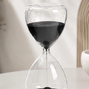 May include: A clear glass hourglass featuring a rounded top and bottom bulb connected by a narrow neck. The interior contains fine black sand, with a portion of the sand currently flowing through the center opening into the bottom glass chamber.