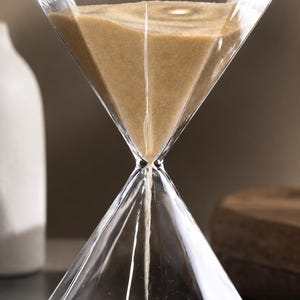 Puede incluir: Un reloj de arena de cristal transparente con forma de diamante facetado, que contiene arena fina de color beige. La sección superior está parcialmente llena de arena, que fluye a través de un punto central estrecho hacia la inferior.