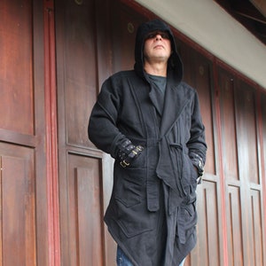 May include: A black hooded jacket with a long, asymmetrical hemline. The jacket has a belt and a zipper closure. The person is wearing blue jeans.