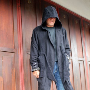 May include: A person wearing a black hooded jacket with a long, flowing design. The jacket has a zipper closure and a belt at the waist. The person is wearing blue jeans and is standing in front of a wooden door.