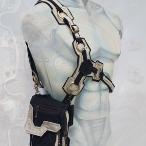 Pode incluir: Uma bolsa transversal de lona preta e branca com um design geom&eacute;trico e v&aacute;rios bolsos. A bolsa &eacute; usada no ombro de um manequim com uma al&ccedil;a branca e preta.