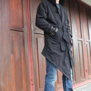 May include: A black hooded jacket with a long, asymmetrical hemline. The jacket has a belt and pockets. The person is wearing blue jeans.