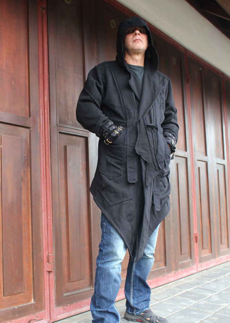 May include: A black hooded jacket with a long, asymmetrical hemline. The jacket has a belt and a zipper closure. The person is wearing blue jeans.