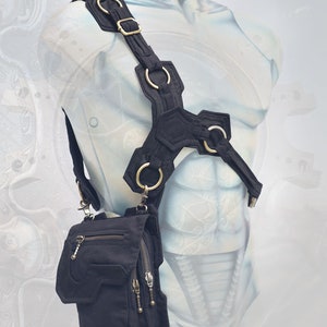 Pode incluir: Uma bolsa de ombro de lona preta com v&aacute;rios bolsos e fechos de correr. A bolsa est&aacute; presa a um arn&ecirc;s preto e prateado com v&aacute;rios an&eacute;is de metal.