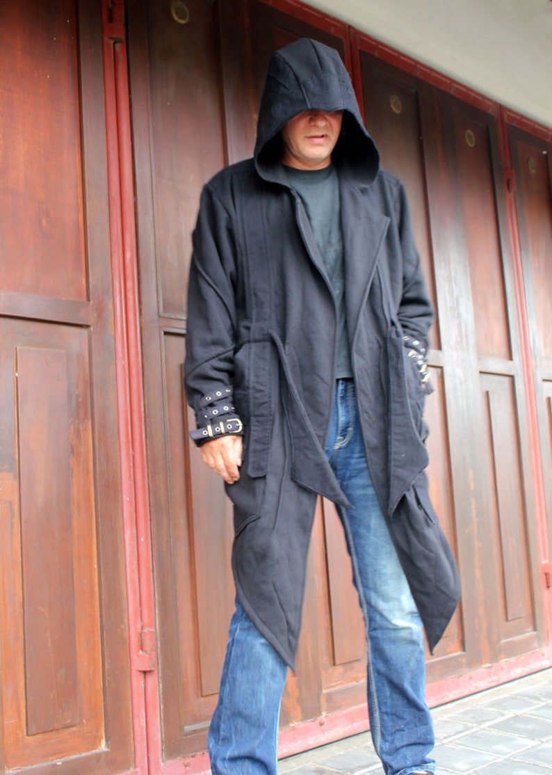 May include: A person wearing a black hooded jacket with a long, flowing design. The jacket has a zipper closure and a belt at the waist. The person is wearing blue jeans and is standing in front of a wooden door.