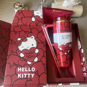 Puede incluir: Un vaso Stanley Quencher H2.0 FlowState de color rojo con un patrón de Hello Kitty y corazones, empaquetado en una caja roja a juego con gráficos de Hello Kitty. El vaso incluye un asa, una tapa transparente y una pajita, con el texto Hello Kitty.