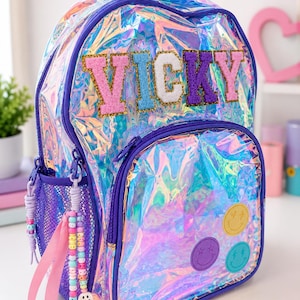 May include: A holographic iridescent backpack with purple trim and a purple mesh side pocket. The name VICKY is displayed in glittery, multi-coloured block letters across the top. Three circular smiley face patches in yellow, purple, and teal are attached to the front pocket, which also features beaded zip pulls.