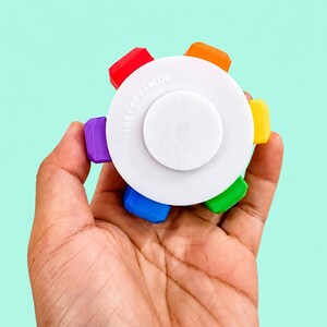 May include: A circular white plastic fidget toy with a central button and six protruding colorful tabs in red, orange, yellow, green, blue, and purple. The text THE POP PARLOR is embossed on the top surface of the white circular base.