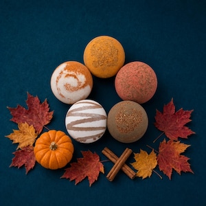 May include: Five spherical bath bombs in shades of orange, white, pink, and brown, arranged in a circular pattern. The set includes a small orange pumpkin and two cinnamon sticks, surrounded by scattered red and yellow maple leaves.