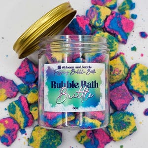 May include: A clear plastic jar filled with multi-colored bath brittle in shades of pink, blue, and yellow. The jar has a gold lid and a label that reads "Fizzing Bubble Bath Bubble Bath Brittle 8oz". Loose pieces of the brittle surround the jar.