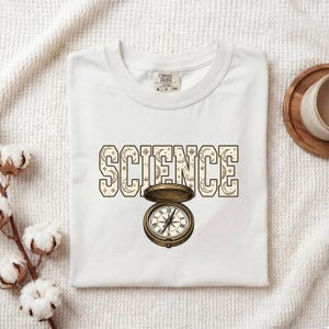 Puede incluir: Una camiseta blanca con la palabra SCIENCE en letras grandes de bloque rellenas con un patrón celestial de lunas y estrellas. Debajo del texto hay una ilustración detallada de una brújula de bolsillo abierta de color latón antiguo.