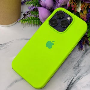 Puede incluir: Una funda para smartphone de color verde lima neón brillante con un logotipo de Apple centrado y un módulo de cámara de tres lentes en la parte posterior. La funda tiene un acabado mate suave y cubre la parte trasera y los lados del dispositivo.