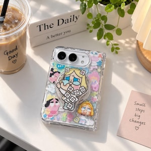 May include: A clear smartphone case featuring a central circular pop-out grip decorated with a cartoon character illustration. The case is filled with various colorful, small, flat character stickers and shapes, including floral and heart motifs.