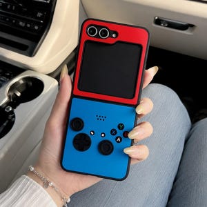 May include: A protective phone case designed to resemble a handheld video game console. The top section is red with a black rectangular cutout for the camera, while the bottom section is blue with black buttons, a D-pad, and joystick controls.