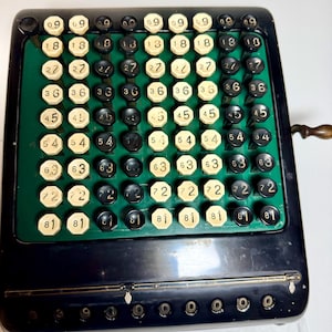 May include: A vintage mechanical adding machine featuring a black metal casing and a green faceplate. The device includes a grid of octagonal keys in alternating black and white colors, each marked with numerical values from 09 to 81.