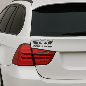 Sticker Grogu bébé à bord noir posé sur l’arrière d’une voiture blanche, près du feu rouge, pour montrer son rendu réel sur carrosserie. Le design Baby Yoda reste visible, propre et discret, avec le texte “BEBE A BORD” en lettres épaisses. Cet autocollant vinyle 20x7 cm convient aux voitures, vitres, gourdes, ordinateurs, portables et supports lisses, parfait comme décoration auto ou idée cadeau originale pour jeunes parents et fans de Star Wars.