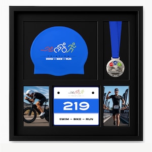 May include: A black shadow box frame containing a blue swim cap with a triathlon logo, a silver medal with a blue ribbon, two photographs of a triathlete, and a white race bib featuring the number 219 and the text SWIM - BIKE - RUN.