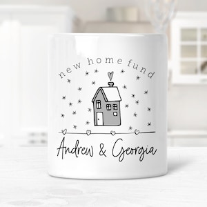May include: White ceramic money box with a black and grey illustration of a house with a chimney and a heart on top. The text "new home fund" is written in a circle around the house. The names "Andrew & Georgia" are written below the house.