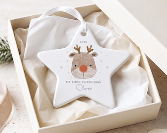 Könnte beinhalten: Ein weißer, sternförmiger Keramikschmuck mit einer Aquarell-Illustration eines Rentiers, dem Text 2026, MY FIRST CHRISTMAS und dem Namen Oliver. Der Anhänger hängt an einem weißen Band in einer offenen cremefarbenen Geschenkbox.