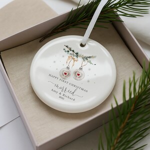 Round white ceramic ornament resting inside an open gift box lined with beige fabric and pine sprigs.
