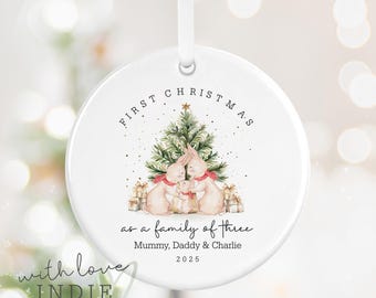 May include: A white ceramic Christmas ornament with a ribbon for hanging. The ornament features a watercolor illustration of a family of three bunnies in front of a Christmas tree. Text reads "First Christmas as a family of three" and "Mummy, Daddy & Charlie 2025."