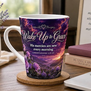 May include: A white ceramic mug featuring a vibrant sunset landscape with purple roses in the foreground. The text reads Wake Up to Grace, His mercies are new every morning, and LAMENTATIONS 3:22-23. The mug has a curved handle and sits on a round cork coaster.