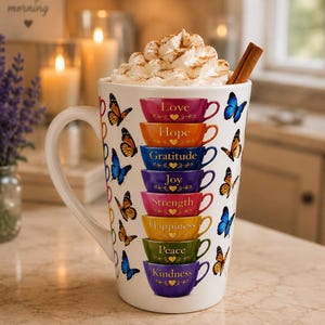 Könnte beinhalten: Ein weißer Keramikbecher gefüllt mit Schlagsahne und einer Zimtstange. Der Becher zeigt einen vertikalen Stapel aus acht bunten Tassen mit den Aufschriften Love, Hope, Gratitude, Joy, Strength, Happiness, Peace und Kindness, umgeben von orangefarbenen und blauen Schmetterlingen.