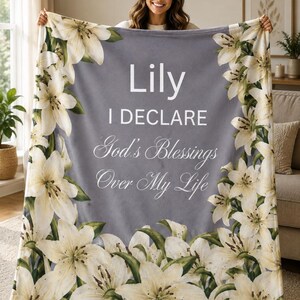 May include: A grey fleece blanket featuring a border of white lily flowers and green leaves. The center text reads: Lily I DECLARE God's Blessings Over My Life. The blanket has a soft texture and is displayed in a vertical orientation with floral patterns.