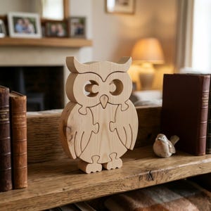 May include: A light-coloured wooden owl puzzle featuring a natural grain finish. The owl is composed of several interlocking pieces that form the shape of the bird, including distinct head, wing, and body sections with visible puzzle cut lines.