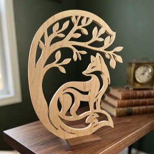May include: A light-coloured wooden cutout sculpture featuring a stylised fox sitting beneath the branches of a tree. The design is circular, with the fox positioned in the lower centre and tree branches arching over it, all mounted on a base.