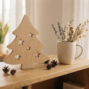 Peut inclure: Une silhouette de sapin de Noël en bois clair découpée avec cinq perforations en forme d'étoile. L'objet est posé sur une surface en bois à côté d'une tasse en céramique beige contenant de la lavande et du blé séchés et des pommes de pin.