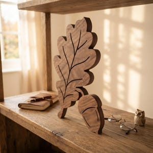 May include: A dark brown wooden cutout shaped like an oak leaf with a matching acorn piece positioned at its base. The leaf features engraved vein details, and the acorn has a carved cap line, both displaying a natural wood grain texture.