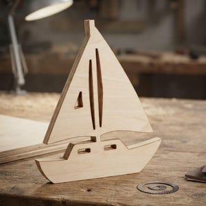 May include: A light-colored wooden sailboat cutout featuring a layered plywood edge design. The sailboat has a triangular sail with two vertical cutouts and a small triangular window, along with a hull featuring two rectangular cutouts.
