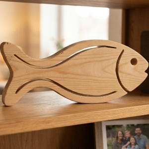 Puede incluir: Una figura de pez de madera de color claro con un acabado de grano natural suave. El diseño incluye un detalle recortado que define la cola, el cuerpo y la cabeza, con un orificio circular que sirve como el ojo del pez.