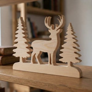 Può includere: Un ornamento in legno chiaro intagliato che raffigura la sagoma di un cervo posizionata tra due alberi sempreverdi stilizzati. L'intero pezzo è montato su una base rettangolare piatta in legno, che mette in risalto le venature naturali.