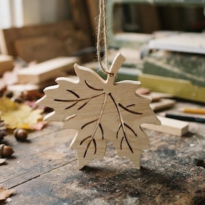 May include: A light-colored wooden maple leaf ornament with laser-cut vein details. The leaf is suspended by a thin, natural-toned jute twine loop attached to the top stem, showcasing a smooth, unfinished wood texture and a distinct botanical shape.