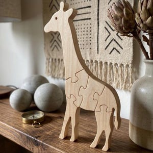 Peut inclure: Une figurine de girafe en bois clair conçue comme un puzzle. La girafe se tient debout, présentant une finition lisse en grain de bois naturel et des segments de puzzle imbriqués qui forment le corps, le cou et les pattes de l'animal.