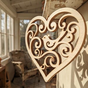 May include: A light-colored wooden heart-shaped ornament featuring intricate laser-cut scrollwork and a central bird silhouette. The decorative piece is suspended by a thin twine string, showcasing layered wood grain edges and a hollow design.