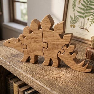 May include: A light brown wooden dinosaur puzzle composed of three interlocking pieces. The figure features a long neck, a head with a small dot for an eye, and a series of plates along its back. The grain of the wood is visible throughout the surface.