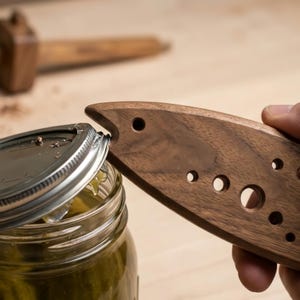 Könnte beinhalten: Ein dunkelbrauner Glasöffner aus Holz in Fischform mit mehreren kreisförmigen Aussparungen unterschiedlicher Größe am Körper. Das Werkzeug ist so angesetzt, dass es den Metalldeckel eines mit grünen Gewürzgurken gefüllten Glases anhebt.