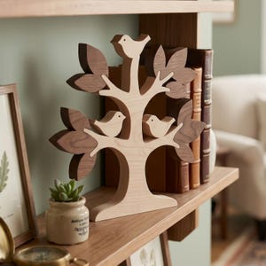 Puede incluir: Una escultura decorativa de árbol de madera con un tronco y ramas de color claro, con tres pájaros tallados posados en las ramas. El árbol está acentuado con hojas de madera de color marrón oscuro unidas a las ramas, creando un contraste bicolor.