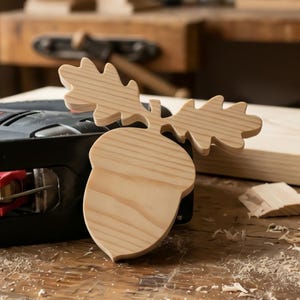 May include: A light-coloured wooden cutout shaped like an acorn with two oak leaves attached to the top. The wood grain is visible across the surface of the piece, which features smooth, rounded edges and a natural, unfinished light tan appearance.
