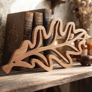 Peut inclure: Une découpe en bois brun clair en forme de feuille de chêne avec une finition lisse et un petit trou circulaire près de la tige. L'objet présente un design ajouré détaillé qui met en valeur le grain naturel et la texture du bois.
