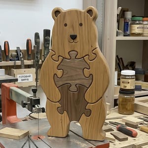 May include: A standing wooden bear puzzle featuring a light brown outer frame and two darker brown, interlocking puzzle pieces in the center. The bear has simple dark brown eyes and a nose, with a smooth finish showing natural wood grain patterns.