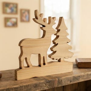 May include: A light-colored wooden cutout featuring the silhouette of a standing deer with antlers positioned next to a pine tree. The object is mounted on a rectangular wooden base and displays natural wood grain patterns with darkened edges.
