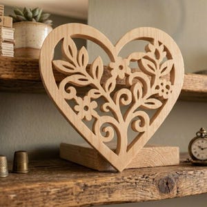 May include: A light-colored wooden heart-shaped cutout featuring an intricate laser-cut design of swirling vines, leaves, and small flowers. The heart is mounted on a rectangular wooden base, showcasing a natural wood grain texture throughout.