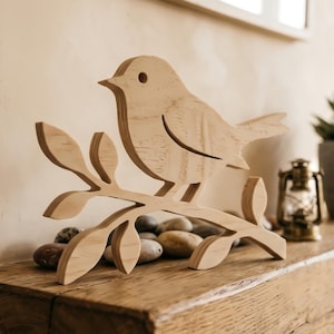 May include: A light-colored wooden cutout sculpture of a bird perched on a branch with several leaves. The bird has a simple circular eye and a pointed beak. The wood grain is visible throughout the surface of the flat, layered-style figure.